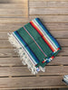 A folded multicolored serape blanket with cream fringe, displayed on a weathered brown wooden deck.
