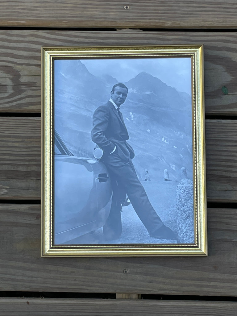 A framed vintage black and white photograph Sean Connery as James Bond in a suit leaning against a car in a mountain setting.