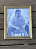 A framed monochrome portrait print of Johnny Cash, shown in a gold-toned frame against a weathered wooden plank background.