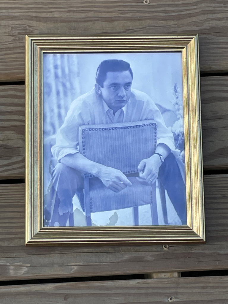 A framed monochrome portrait print of Johnny Cash, shown in a gold-toned frame against a weathered wooden plank background.