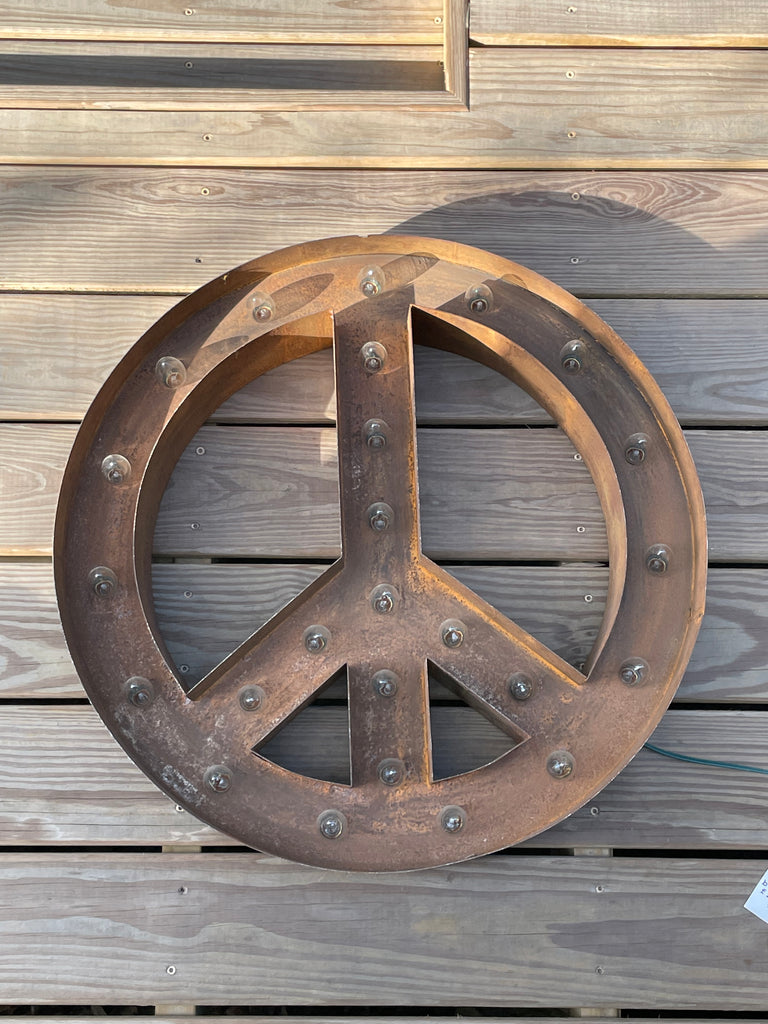 A large (24") circular metal wall sign featuring a peace symbol, finished in a rustic weathered bronze-brown color with rivet details, mounted on a wooden deck.