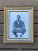 A framed black and white portrait photograph of a Paul Newman, set in an ornate gold frame against a dark wood plank background.