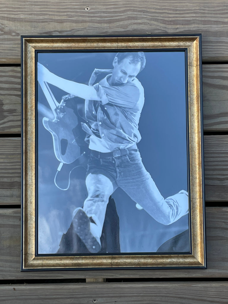 A framed black and white photo print of a guitarist, Pete Townsend, in motion, mounted on a dark wood plank background. The print features a blue tint and is enclosed in a double-matted frame with a gold inner border and a black outer frame.