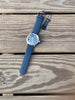 A wristwatch with a blue strap and a silver-toned round case, laid diagonally on a rustic wooden plank surface. The watch has a blue dial and a black hangtag attached. Made by Shinola, Detroit.