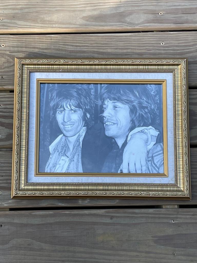 A framed monochrome portrait print of Rolling Stones members Mick and Keith, set in an ornate gold frame and mounted against a weathered wooden plank wall.