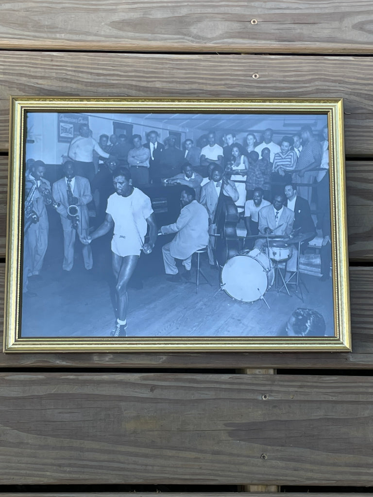 A framed black and white photograph of a group of people, including jazz band on hand for Sugar Ray Robinson's workout. 1949.