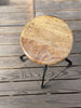 A rustic round wooden stool with a distressed brown and beige seat, supported by a black metal frame, set on a weathered gray wooden deck.