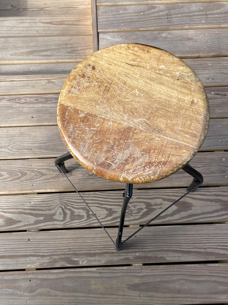 A rustic round wooden stool with a distressed brown and beige seat, supported by a black metal frame, set on a weathered gray wooden deck.