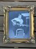 An ornate gold picture frame holding a blue-toned black-and-white photograph of Elvis playing an acoustic guitar, displayed against a vertical wood plank background.